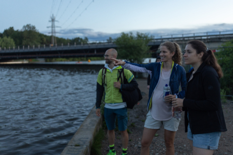Drei Personen stehen in der Abenddämmerung an einem Flussuferweg, zwei Frauen und ein Mann, alle in sportlicher Kleidung. Eine Frau zeigt auf das Wasser, während die anderen in die gleiche Richtung schauen. Im Hintergrund sind eine Brücke und Bäume zu sehen.