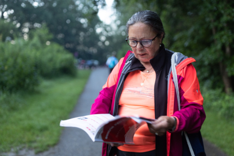 Eine ältere Frau mit Brille und rosa Jacke steht auf einem Weg im Freien und blättert in einer Zeitschrift oder einem Heft. Im Hintergrund sind üppige grüne Bäume und Gras zu sehen.