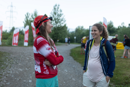 Zwei Frauen stehen im Freien auf einem Kiesweg, lächeln und unterhalten sich. Die eine trägt ein rotes Sportoutfit und eine Mütze, die andere, sichtlich schwanger, eine blaue Jacke und beigefarbene Shorts. Im Hintergrund sind Fahnen und andere Menschen zu sehen.