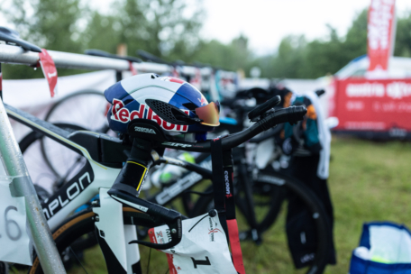 Nahaufnahme eines Rennrads mit einem Red Bull-Helm auf dem Lenker, das in einer Wechselzone bei einer Radveranstaltung im Freien abgestellt ist. Im Hintergrund sind andere Fahrräder und Grünflächen zu sehen.