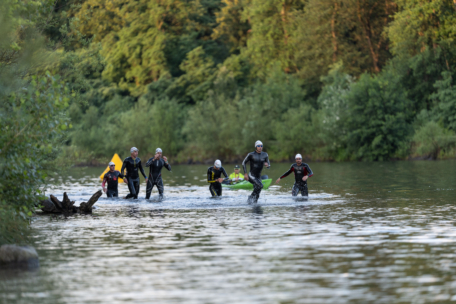 Eine Gruppe von Menschen in Neoprenanzügen und Badekappen läuft durch einen seichten Fluss, der von dichten grünen Bäumen umgeben ist, und nimmt an einem Schwimmwettbewerb im Freien teil.