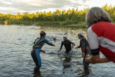 Mehrere Personen in Neoprenanzügen helfen sich gegenseitig im flachen Wasser eines Sees in der Nähe eines grünen, von Bäumen gesäumten Ufers, wobei eine Person ihre Hand ausstreckt und eine andere Person im Vordergrund zusieht.