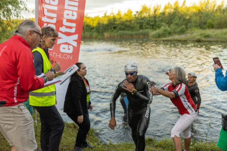 Ein Athlet im Neoprenanzug verlässt während eines Triathlons einen See. Er wird von Freiwilligen und Veranstaltungsmitarbeitern mit Klemmbrettern und Warnwesten begrüßt, während andere in der Nähe eines straXtreme-Banners am Ufer zusehen.