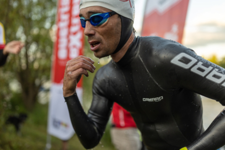 Ein Triathlet in einem schwarzen Neoprenanzug, einer weißen Badekappe und einer blauen Schwimmbrille taucht aus dem Wasser auf und wirkt konzentriert und entschlossen, während im Hintergrund unscharfe Veranstaltungsbanner und Grünpflanzen zu sehen sind.