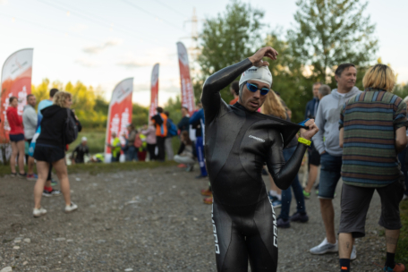 Ein Schwimmer in Neoprenanzug und Badekappe passt seinen Anzug bei einer Veranstaltung im Freien an, umgeben von Menschen, Bannern und Grünflächen. Es herrscht eine lebhafte Atmosphäre mit Zuschauern und Teilnehmern.