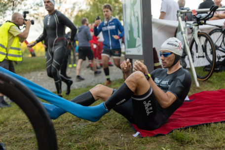 Ein Triathlet in Neoprenanzug und Badekappe sitzt auf einem roten Handtuch und zieht sich blaue Socken über die Füße, während andere Athleten und ein Fotograf im Hintergrund in der Wechselzone des Rennens zu sehen sind.