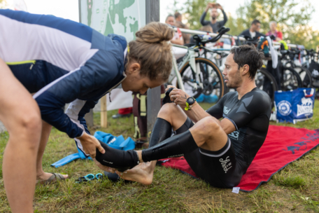Ein Mann in einem Neoprenanzug sitzt auf einem roten Handtuch und bereitet sich auf einen Triathlon vor. Eine Frau hilft ihm, sein Bein im Neoprenanzug anzupassen. Im Hintergrund sind Fahrräder und andere Sportler zu sehen.