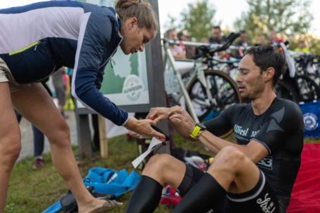 Eine Frau hilft einem männlichen Triathleten, der auf der Wiese sitzt, beim Anlegen eines Armbandes. Im Hintergrund sind Fahrräder geparkt, und andere Personen stehen in der Nähe. Der Sportler sieht müde und konzentriert aus.