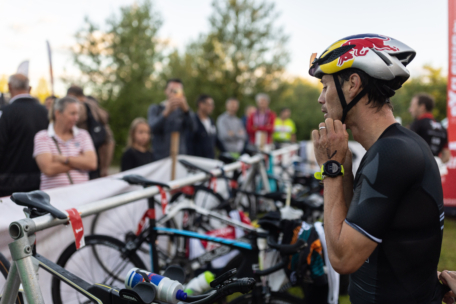 Ein männlicher Triathlet, der einen Helm und eine Sportausrüstung trägt, passt seinen Gurt in der Nähe einer Reihe von geparkten Fahrrädern in der Wechselzone an, mit Zuschauern und Bäumen im Hintergrund.