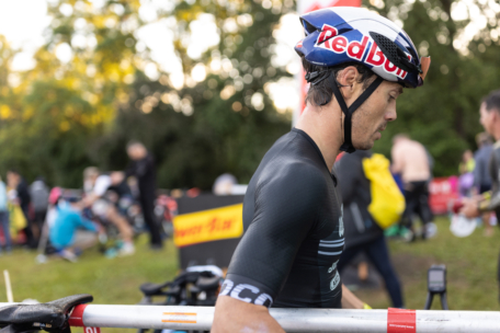 Ein Radfahrer mit Red-Bull-Helm und schwarzer Ausrüstung trägt ein Fahrrad auf der Schulter während einer Veranstaltung im Freien, mit unscharfen Teilnehmern und Grünflächen im Hintergrund.