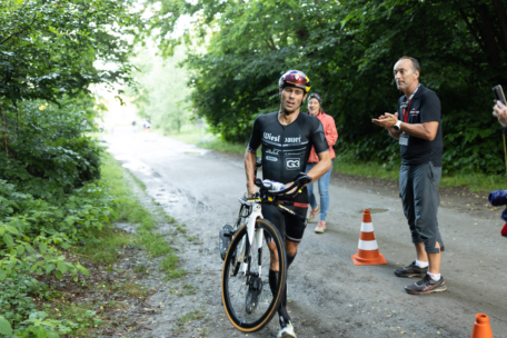 Ein Radfahrer in Sportkleidung fährt mit seinem Fahrrad einen Waldweg entlang, vorbei an einem orangefarbenen Verkehrskegel. Zwei Menschen stehen in der Nähe, einer klatscht, der andere macht ein Foto, während auf beiden Seiten des Weges Grünflächen zu sehen sind.
