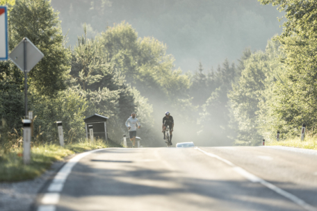 Eine neblige Morgenszene auf einer von üppigem Grün umgebenen Landstraße, die einen Radfahrer zeigt, der bergauf fährt, während eine Person in der Nähe joggt. Das Sonnenlicht fällt durch die Bäume und schafft eine friedliche Atmosphäre.