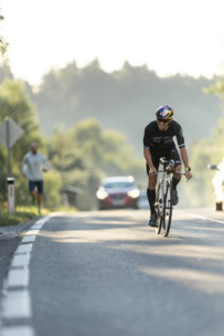 Ein Radfahrer mit Helm und schwarzem Outfit fährt auf einer sonnenbeschienenen Straße, während im Hintergrund ein Auto und eine Person joggen. Die Szene ist von Grünflächen und Bäumen umgeben.