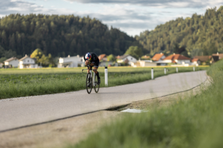 Ein Radfahrer in Rennkleidung fährt auf einem Rennrad auf einem gewundenen, von grünem Gras gesäumten ländlichen Weg, mit einem Dorf und bewaldeten Hügeln im Hintergrund unter einem teilweise bewölkten Himmel.