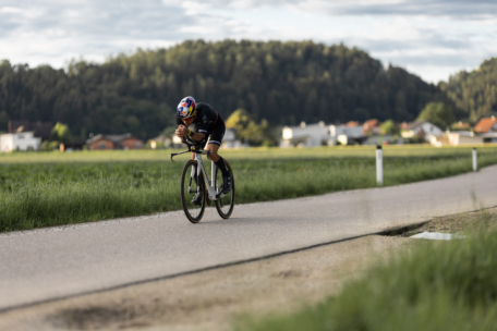 Ein Radfahrer mit Helm fährt mit seinem Rennrad auf einer Landstraße, umgeben von grünen Feldern, mit Bäumen und Häusern im Hintergrund unter einem bewölkten Himmel.
