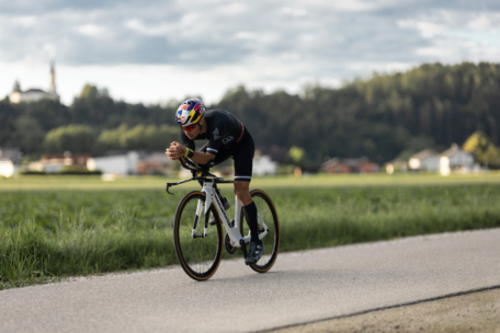 Ein Radfahrer in dunkler Rennkleidung und mit Helm fährt mit seinem Rennrad auf einem gepflasterten Weg durch eine ländliche Gegend, mit grünem Gras und unscharfen Bäumen und Gebäuden im Hintergrund.