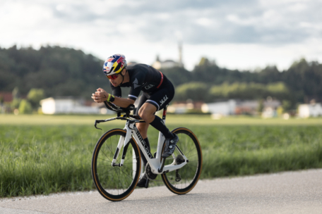 Ein Radfahrer in schwarzer Kleidung und mit Helm fährt auf einem weißen Rennrad auf einer gepflasterten Straße neben einem grasbewachsenen Feld, mit Bäumen und einem verschwommenen Gebäude im Hintergrund unter einem bewölkten Himmel.