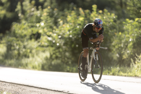 Ein Radfahrer in schwarzer Kleidung und Helm fährt auf einem weißen Fahrrad auf einer sonnenbeschienenen Straße, umgeben von grünem Laub, und lehnt sich in einer aerodynamischen Position nach vorne.