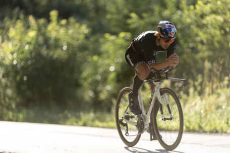 Ein Radfahrer in Rennkleidung und Helm fährt mit einem aerodynamischen Rennrad auf einer sonnenbeschienenen Straße, umgeben von grünem Laub im Hintergrund.