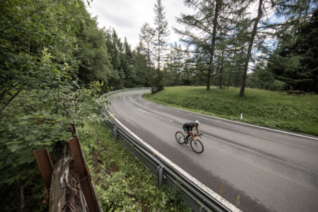 Ein Radfahrer fährt schnell um eine scharfe, leere Kurve auf einer gepflasterten Straße, umgeben von hohen Bäumen und üppigem Grün unter einem bewölkten Himmel.