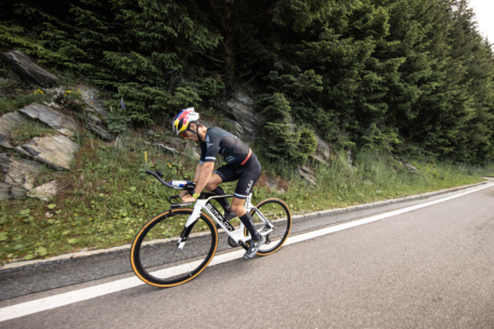 Ein Radfahrer, der einen Helm und eine Rennausrüstung trägt, fährt mit seinem Rennrad bergauf auf einer asphaltierten Straße neben einem dichten Wald und einem felsigen Abhang.