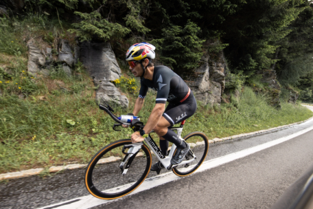 Ein Radfahrer in schwarzer Kleidung und mit Helm fährt mit seinem Rennrad bergauf auf einer kurvenreichen Straße, die von Felsen und Grünflächen gesäumt ist.