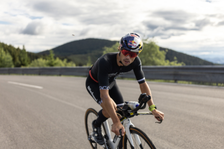 Ein Radfahrer mit schwarzem Trikot, Sonnenbrille und Red-Bull-Helm fährt mit seinem Rennrad auf einer offenen Landstraße mit grünen Hügeln und einem bewölkten Himmel im Hintergrund.