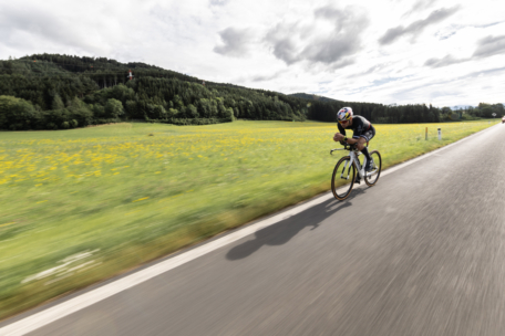 Ein Radfahrer fährt schnell auf einer Straße durch die Landschaft, mit grünen Feldern und Bäumen auf beiden Seiten und Hügeln im Hintergrund unter einem teilweise bewölkten Himmel.