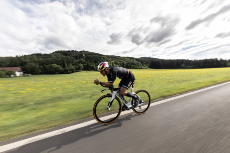Ein Radfahrer in Rennkleidung fährt mit seinem Rennrad schnell eine Landstraße entlang, mit grünen Feldern und bewaldeten Hügeln im Hintergrund unter einem teilweise bewölkten Himmel.