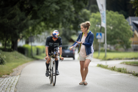 Ein Radfahrer in Rennkleidung fährt auf einer Straße, während eine Frau in Freizeitkleidung neben ihm geht und mit der Hand gestikuliert. Im Hintergrund sind Bäume und ein Auto zu sehen.