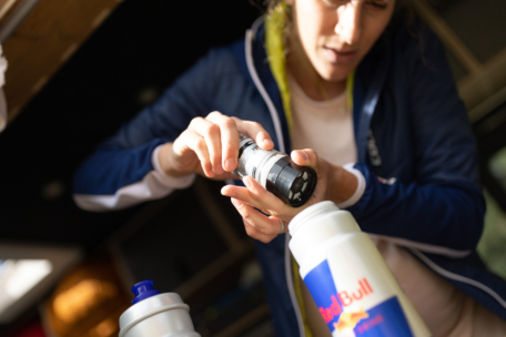 Eine Person in einer blauen Jacke bereitet ein Getränk zu, indem sie Pulver aus einem kleinen Behälter in eine Sportwasserflasche mit der Aufschrift Red Bull einfüllt. Der Fokus liegt auf ihren Händen und der Flasche.