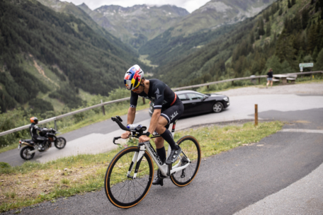 Ein Radfahrer mit Helm und schwarzer Sportkleidung fährt auf einer Straße in einer bergigen Landschaft bergauf, mit einem geparkten Auto, einem Motorradfahrer und üppigen grünen Hügeln im Hintergrund.