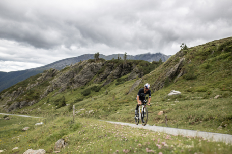 Ein Radfahrer fährt mit seinem Rennrad auf einem schmalen Weg durch eine grasbewachsene, bergige Landschaft unter einem bewölkten Himmel, umgeben von felsigen Hügeln und Wildblumen.