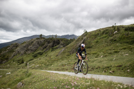 Ein Radfahrer, der einen Helm trägt, fährt mit seinem Rennrad auf einer asphaltierten Bergstraße bergauf, umgeben von grünen Hügeln und felsigem Gelände unter einem bewölkten Himmel.