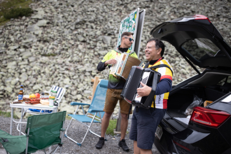Zwei Männer spielen Akkordeon neben einem offenen Autokofferraum am felsigen Straßenrand. Ein kleiner Tisch mit Essen, Getränken und Klappstühlen ist in der Nähe aufgestellt und schafft eine zwanglose Atmosphäre für ein Treffen im Freien.