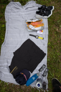 Ein gestreiftes graues Handtuch auf dem Rasen, dazu Laufschuhe, Sandalen, eine Uhr, schwarze Shorts, eine blaue Flasche, zwei Gelpacks, eine Kappe und eine Wasserflasche, alles fein säuberlich darauf angeordnet.