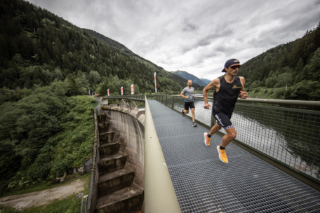 Zwei Männer laufen auf einer Metallbrücke über einen Fluss, umgeben von üppigen grünen Wäldern und Bergen unter einem bewölkten Himmel. Der Läufer vor ihnen trägt ein schwarzes Tank-Top, Shorts und eine Sonnenbrille.