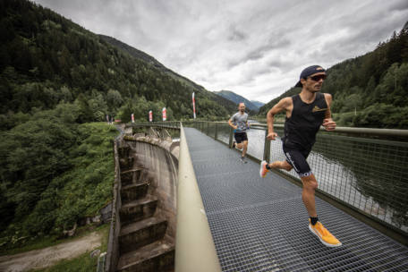 Zwei Männer laufen über eine Brücke über einen Damm, umgeben von üppigen grünen Wäldern und Bergen unter einem bewölkten Himmel. Der vordere Läufer trägt ein sportliches Outfit, eine Sonnenbrille und eine Mütze, während der zweite hinterherläuft.