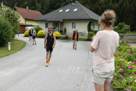 Eine Frau in kurzen Hosen steht in einer Wohnstraße und fotografiert einen Mann in Sportkleidung, der auf sie zugeht. Im Hintergrund laufen andere Menschen in der Nähe von Häusern und Grünanlagen.