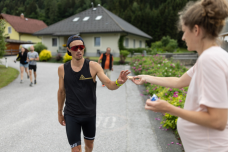 Ein männlicher Läufer in Sportkleidung nimmt während eines Rennens auf einer Wohnstraße einen Gegenstand von einer schwangeren Frau entgegen, während im Hintergrund andere Menschen und Grünflächen zu sehen sind.