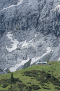 Ein kleines Gebäude steht auf einem grünen Hügel, umgeben von Büschen und Bäumen, mit einer steilen, felsigen Bergwand und Schneeflecken im Hintergrund.
