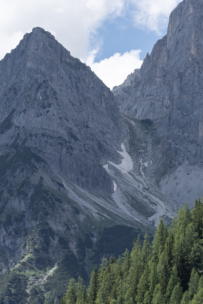 Steile, felsige Berggipfel mit Schneeflecken und grünen Nadelbäumen im Vordergrund unter einem teilweise bewölkten Himmel.