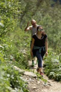Zwei Personen wandern auf einem schmalen, unbefestigten Weg durch eine üppige, grüne Vegetation, wobei die Frau mit Sonnenbrille und Sportkleidung vorausgeht und der Mann hinterherläuft, beide wirken konzentriert und aktiv.