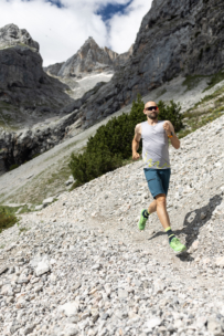 Ein Mann, der eine Sonnenbrille, ein ärmelloses Hemd und kurze Hosen trägt, läuft auf einem felsigen Bergpfad, der von steilen Klippen und Grünflächen umgeben ist, unter einem teilweise bewölkten Himmel.