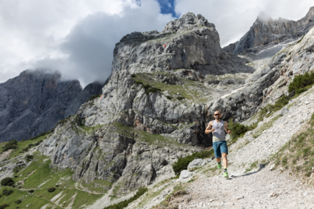 Ein Mann in Sportkleidung läuft auf einem felsigen Bergpfad mit dramatischen Klippen und Wolken im Hintergrund. Die Szene ist hell und im Freien und hebt das zerklüftete Gelände und die beeindruckenden Landschaften hervor.