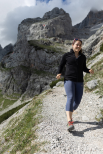 Eine Frau in schwarzer Jacke, grauen Leggings und roter Sonnenbrille joggt auf einem felsigen Bergpfad mit steilen Klippen und grünen Flecken im Hintergrund unter einem teilweise bewölkten Himmel.