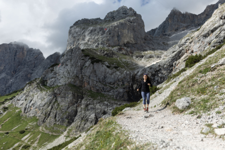 Eine Frau joggt auf einem felsigen Bergpfad, umgeben von steilen Felsen und grünen Flecken, unter einem teilweise bewölkten Himmel.