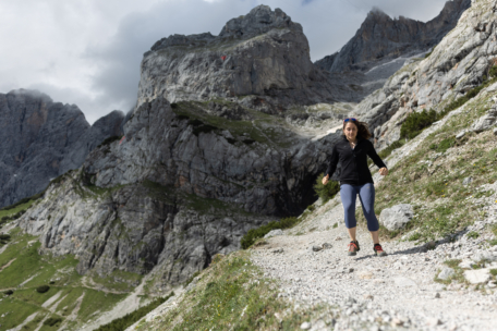 Eine Frau in Sportkleidung läuft auf einem felsigen Bergpfad, umgeben von steilen, schroffen Felsen und bewölktem Himmel.