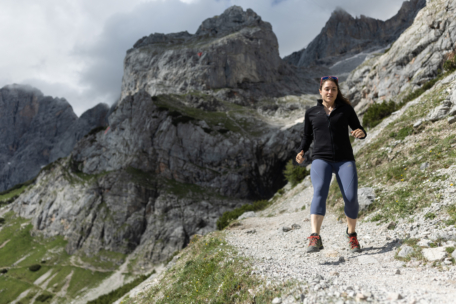 Eine Frau in sportlicher Kleidung läuft auf einem felsigen Bergpfad, umgeben von steilen Felsen und grünen Hängen, mit dramatischen Wolken über dem Kopf.