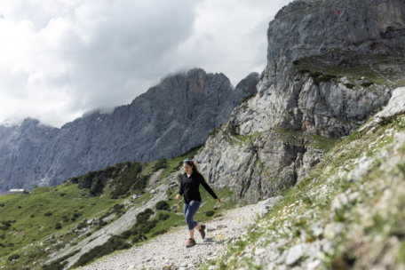 Eine Frau in sportlicher Kleidung läuft auf einem felsigen Bergpfad bergab, umgeben von steilen Felsen und grüner Vegetation unter einem bewölkten Himmel.